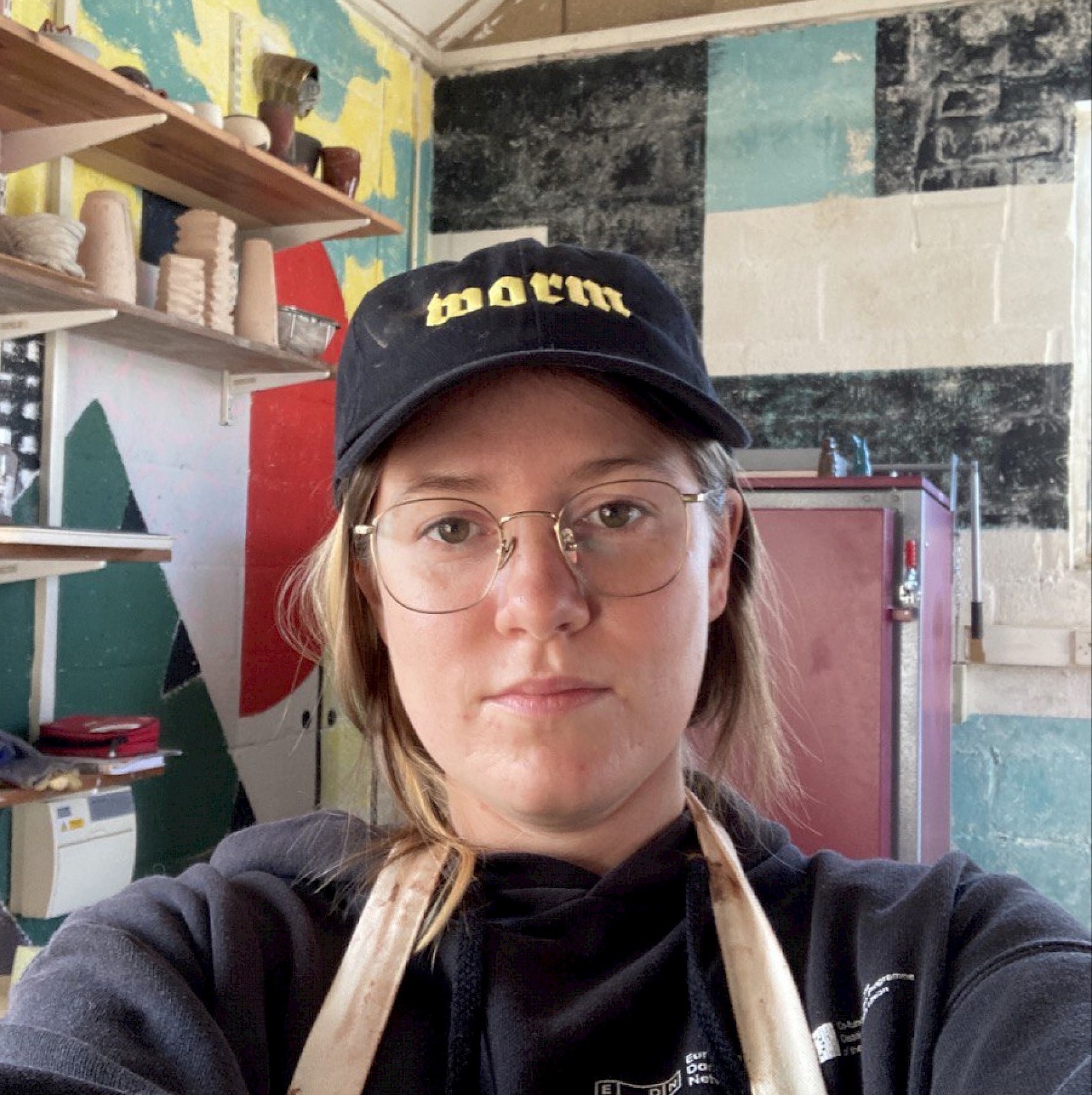 Image of Frances Breden, a white woman with long brown hair, wearing a black baseball cap that says "worm", a black hoodie, and an apron in a colourful ceramics studio