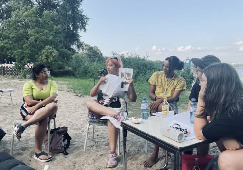 Image of Alungo Xatan, a brown-skinned person with orange hair, talking into a microphone to a circle of listeners.  They are all seated outdoors at a lake next to the water.