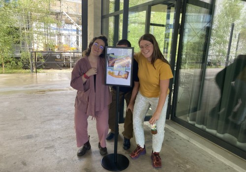 Three people stand in from of a gallery with their arms around each other. Two are stooping down and smiling, and one's face is covered by a sign that says 