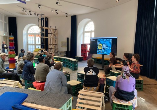 Image of a group sitting down in an art gallery watching three people give a presentation.  There is a cartoon minion on the screen of the presentation.