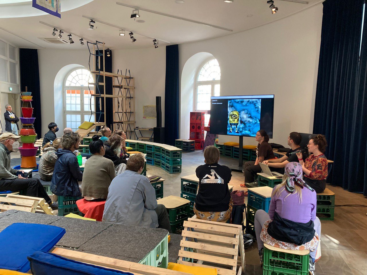 Image of a group sitting down in an art gallery watching three people give a presentation. There is a cartoon minion on the screen of the presentation.