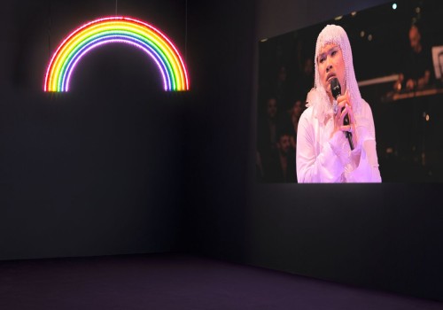 Image of a dark room in a gallery with a neon rainbow sign and a film by the artist Ming Wong where he is singing and wearing a dress.