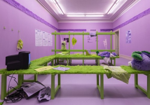 Image of a purple room that is decorated to look like a locker room. There is a green wrap-around bench that is covered in fur. Bright lights shine above.