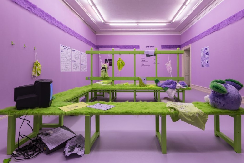 Image of a purple room that is decorated to look like a locker room. There is a green wrap-around bench that is covered in fur. Bright lights shine above.