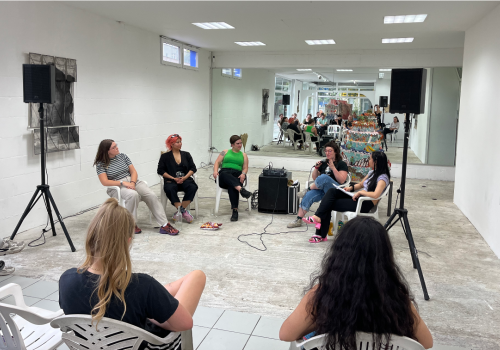 Five people sit in a white gallery giving a talk.  There are speakers and microphones.  Two audience members are facing them.  Their are wearing summery clothing.