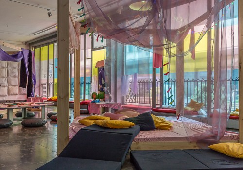 A large white gallery has colourful foils on the window, making the light coloured.  There are beds built of wood and purple fabric and blue mattresses and yellow pillows.  Pillows and favbric hang from the walls.