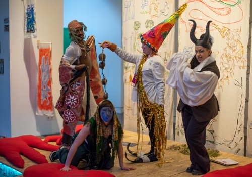 Image of four people on a colourful stage in a gallery. They are wearing oversized princess hats, devils horns, handpainted capes and masks, and surrounded by hand-sewn red cushions and paintings with medieval themes