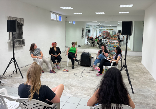Five people sit in a white gallery giving a talk.  There are speakers and microphones.  Two audience members are facing them.  Their are wearing summery clothing.