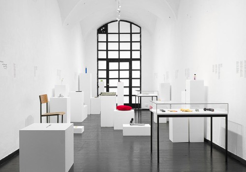 A white gallery has many assorted objects on white plinths, such as a chair and a pillow.