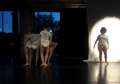 Photo of multiple dancers on a stage with a spotlight. They are all wearing white shirts and diapers.