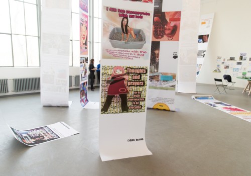 Image of long scrolls of paper 1 meter wide hanging from the ceiling in a white gallery with images of memes from the internet printed on them.