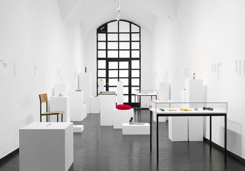 A white gallery has many assorted objects on white plinths, such as a chair and a pillow.