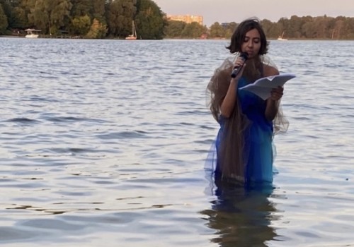 A person wears a blue and brown flowy dress and reads from a book with a microphone while standing in a lake at sunset.