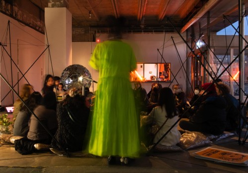 Image of people sitting in a darkly lit gallery in a circle.  A person in a green dress is blurrily walking around the circle.