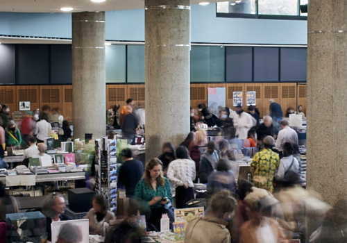 Image of a large crowd of people in an auditorium with cement pillars and many stands with books.