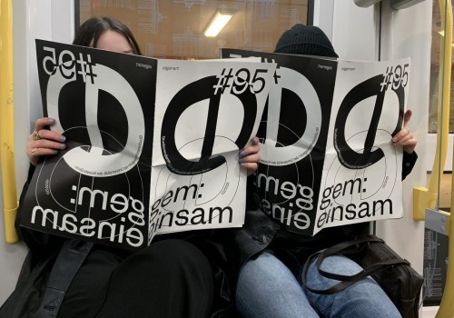 Two people sit in the U-Bahn.  Their faces are covered by large newspapers in black and white saying 