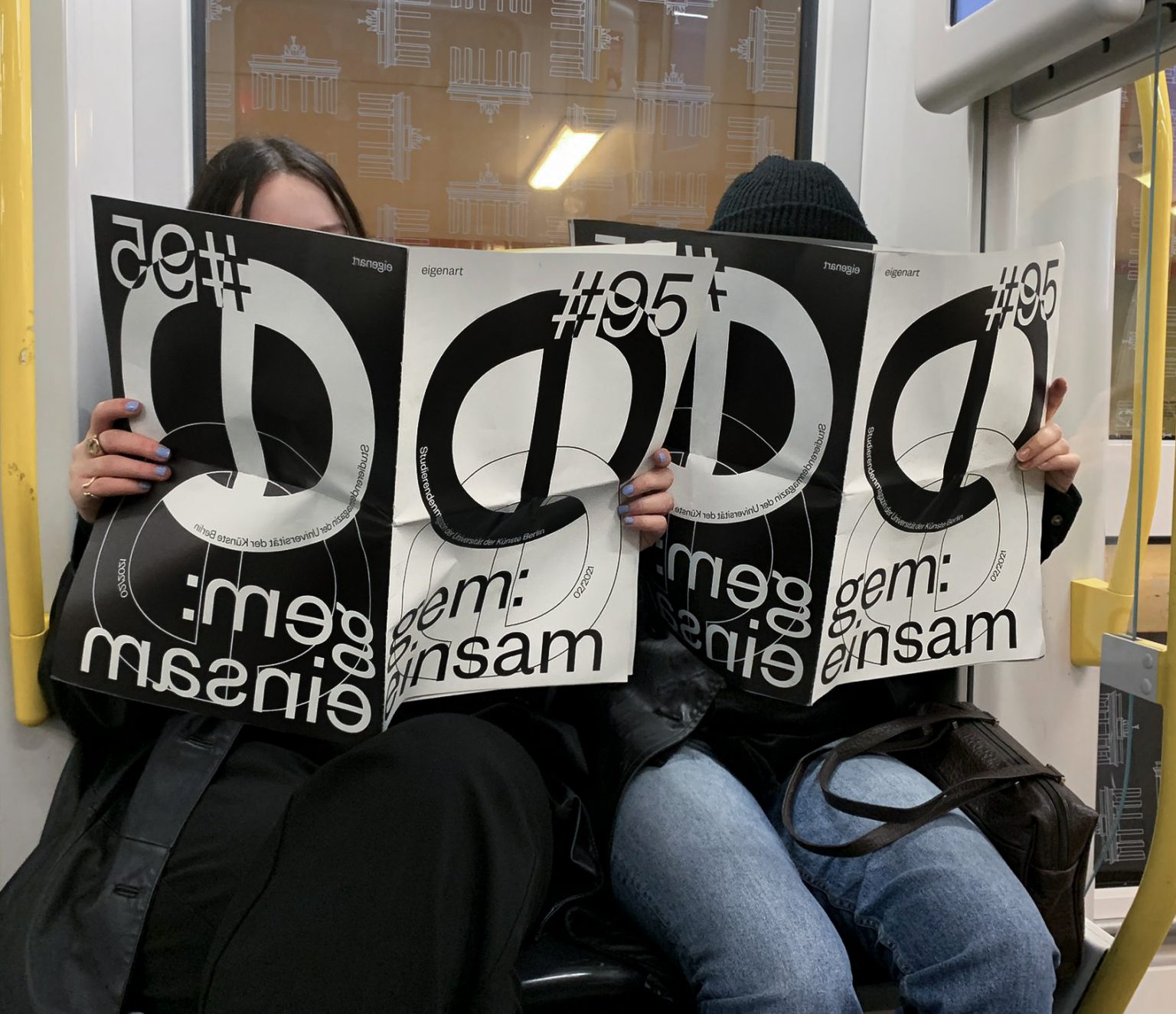 Two people sit in the U-Bahn.  Their faces are covered by large newspapers in black and white saying 