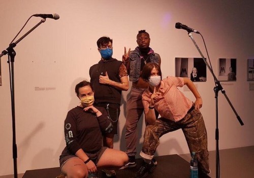 Four people pose on stage, wearing COVID masks and making peace signs with their hands.  There are microhpones around them.