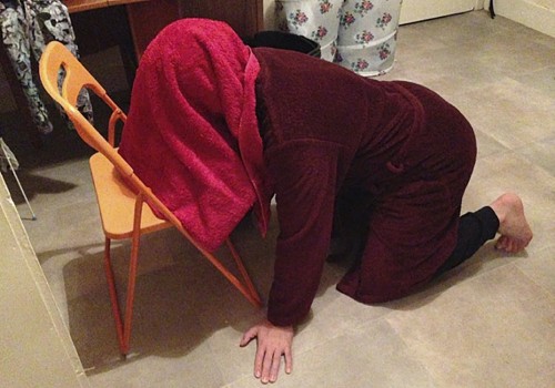 Photo of a person on all fours with their head under a red towel.  Their head and towl are facing the seat of an orange fold-out chair.