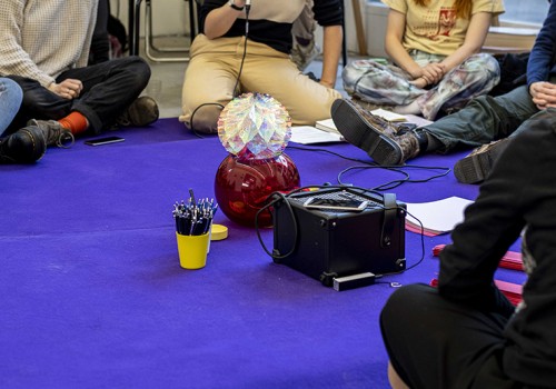 Image of a purple carpet with people's legs sitting on it.  There is a microphone, paper, pens, an amplifier, and a red vase with a colourful paper oragami.