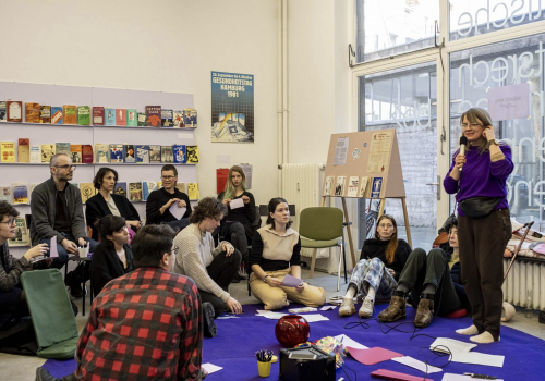 Image of a room full of people, one of them is standing and the rest are sitting. There are books on the wall on shelves, and the standing person appears to be talking.