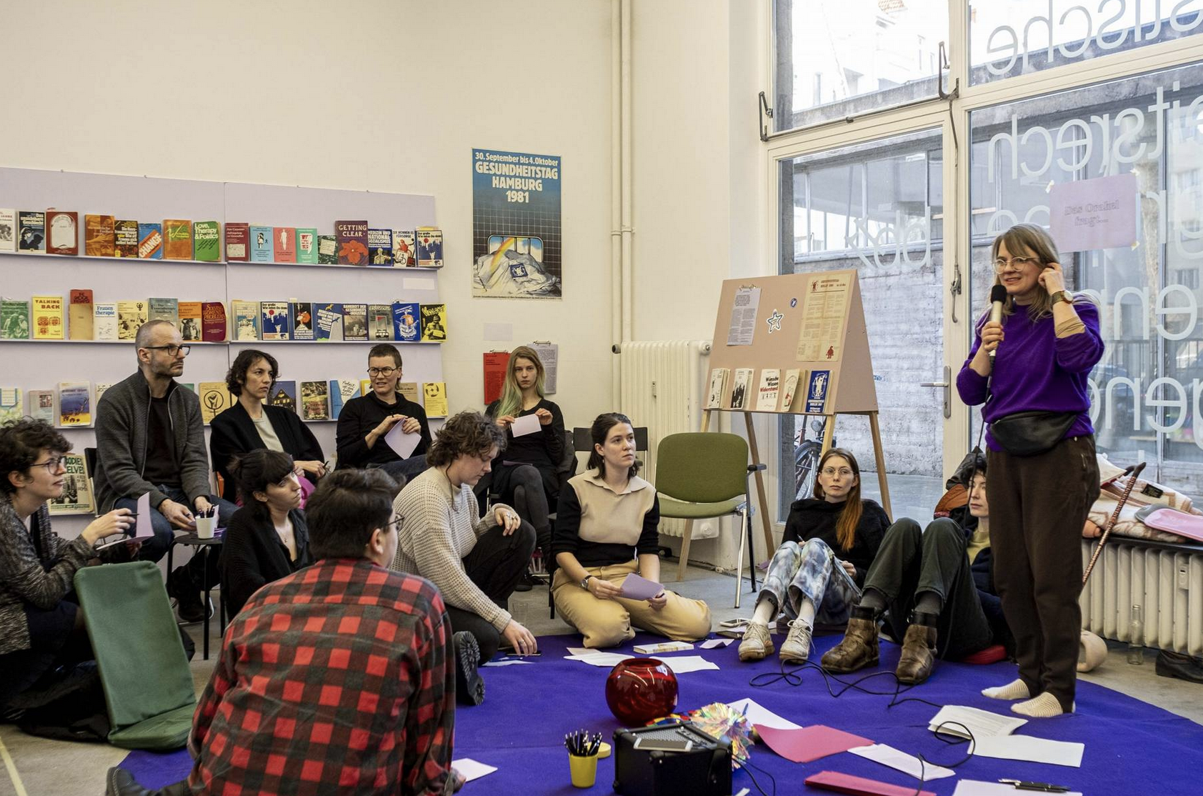 Image of a room full of people, one of them is standing and the rest are sitting. There are books on the wall on shelves, and the standing person appears to be talking.