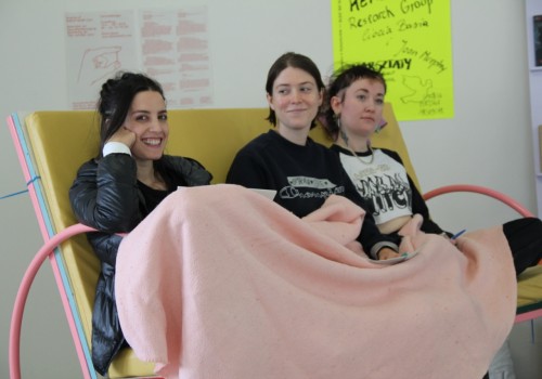 Three people sit in a bench in a gallery with posters on the wall.  They are under a pink planket and smiling.