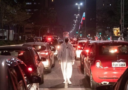 A person wearing a white protective medical suit walks at night on the road past rows of backed-up traffic.